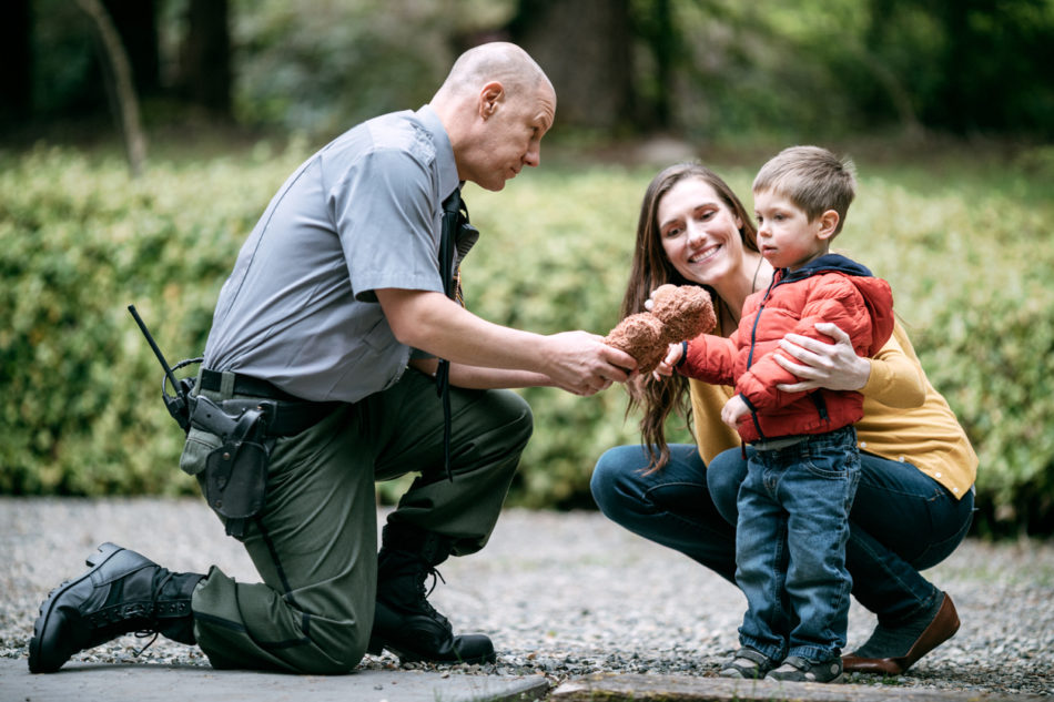 An on duty law enforcement officer talks to a young toddler boy and his mother, giving the child a stuffed a bear