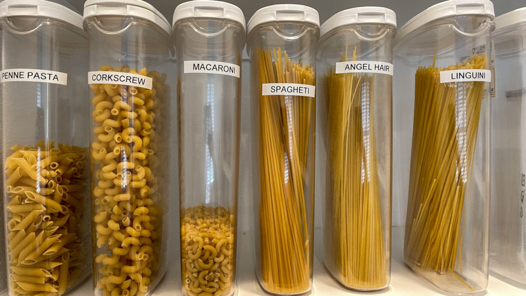 An organized pantry shelf with various types of pasta in plastic containers.