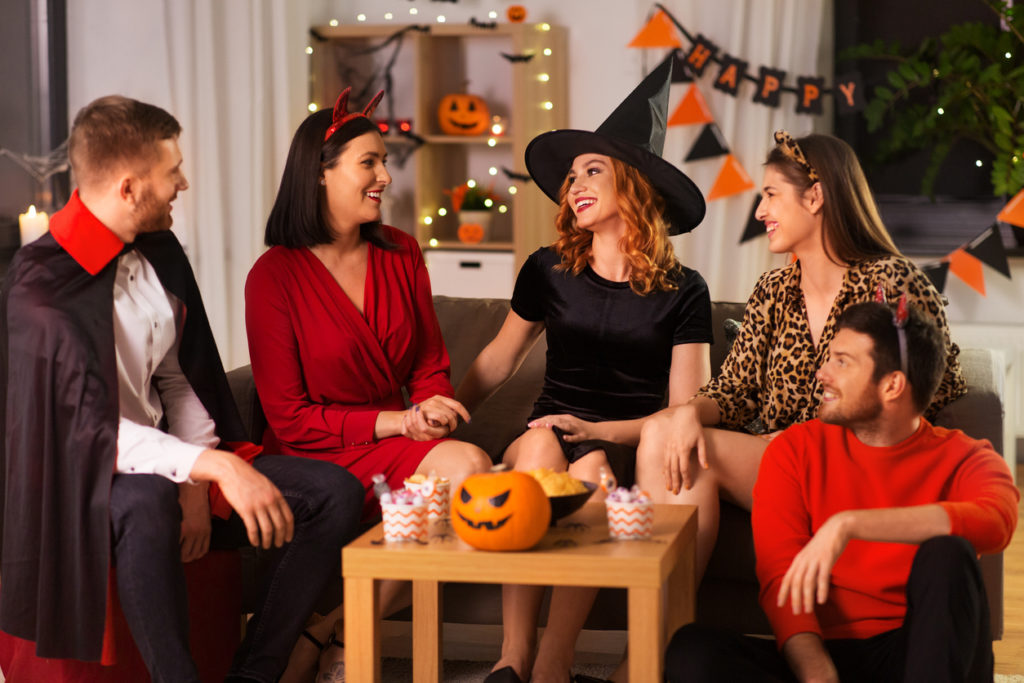 friendship, holiday and people concept - group of happy smiling friends in halloween costumes of vampire, devil, witch and cheetah at home party at night